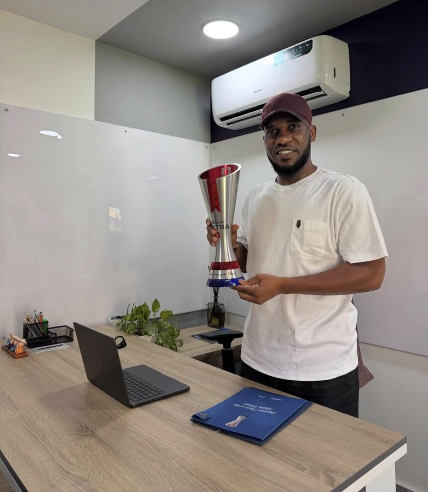 Man in a white t shirt and maroon cap holds a tall silver trophy in a modern office setting  trybalnews