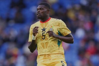 Soccer player in a yellow Ghana kit with number 5, running on a field with blurred blue seats in the background.