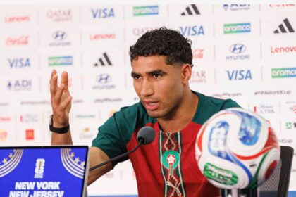 Male footballer at a press conference, speaking into a microphone, with a soccer ball on the table and a branded backdrop behind him.