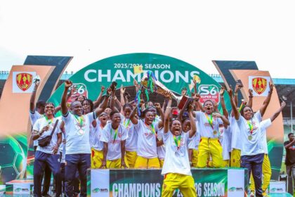 Group of young athletes on a podium celebrate winning the 2025/26 season, raising medals and a trophy amid a champions backdrop.