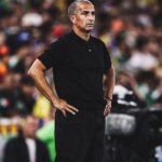 Man in a black polo shirt stands with hands on hips on a soccer field blurred stadium crowd in the background  trybalnews