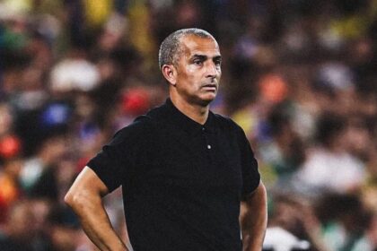Man in a black polo shirt stands with hands on hips on a soccer field, blurred stadium crowd in the background.