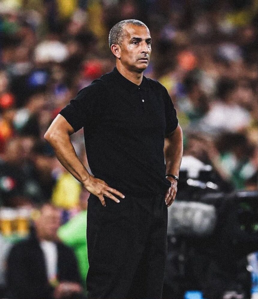 Man in a black polo shirt stands with hands on hips on a soccer field blurred stadium crowd in the background  trybalnews