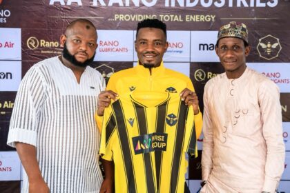 Three men pose together on a promotional backdrop; the center man in a yellow and black jersey holds up a matching kit for display.