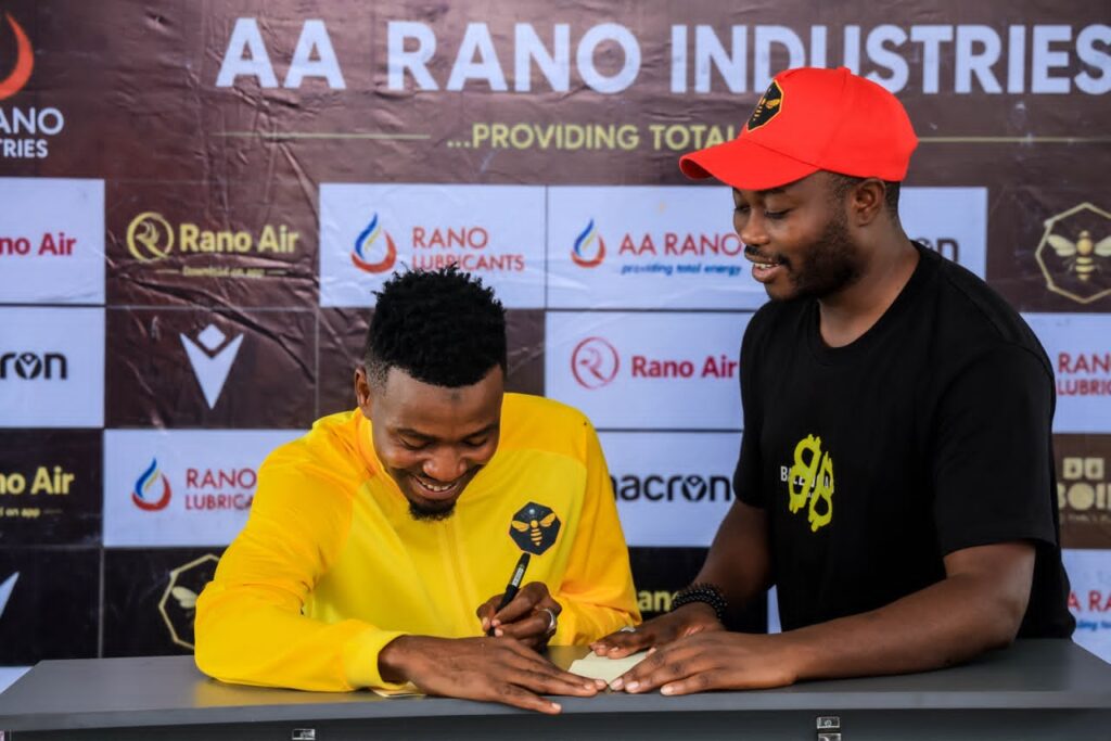 WhatsApp Image 20260622 at 165347 Trybal News Two men at a signing ceremony one in a bright yellow jacket signs a document at a table while the other in a red cap watches trybalnews