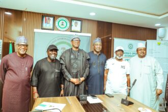 Group of six men in traditional Nigerian attire pose for a photo at a press conference, standing behind a wooden table with microphones and banners in a formal room.