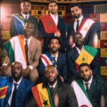 Group of men in suits wearing national flag sashes posing for a formal portrait in front of crates and a banner reading THIS IS AFRICA  trybalnews