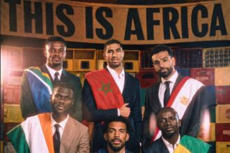 Group of men in suits wearing national flag sashes, posing for a formal portrait in front of crates and a banner reading 'THIS IS AFRICA'.