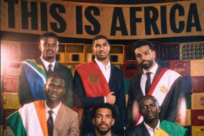 Group of men in suits wearing national flag sashes, posing for a formal portrait in front of crates and a banner reading 'THIS IS AFRICA'.