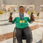Person in a wheelchair in a marble lobby proudly holding two bronze cat statues and wearing two gold medals around the neck the medals hanging over a green and white jacketWhen the semicolon uses  trybalnews