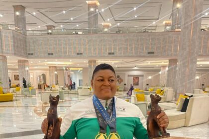 Person in a wheelchair in a marble lobby proudly holding two bronze cat statues and wearing two gold medals around the neck the medals hanging over a green and white jacket.When the semicolon uses?