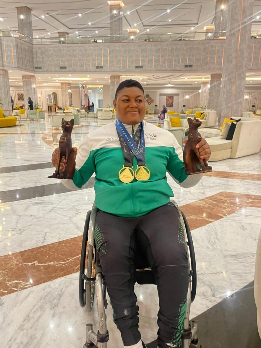 Person in a wheelchair in a marble lobby proudly holding two bronze cat statues and wearing two gold medals around the neck the medals hanging over a green and white jacketWhen the semicolon uses  trybalnews