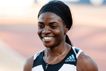 Smiling female sprinter on a track, wearing a black-and-white Adidas top, holding flowers after a race; bib shows 'Amusan' and 'Continental Tour Gold'.