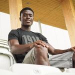 Young man sitting in empty stadium seats smiling at the camera wearing a black t shirt and gray shorts with a wooden roof behind him  trybalnews