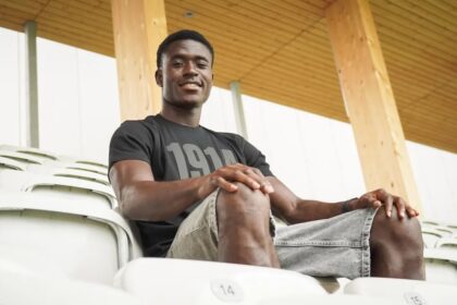 Young man sitting in empty stadium seats, smiling at the camera, wearing a black t-shirt and gray shorts with a wooden roof behind him.