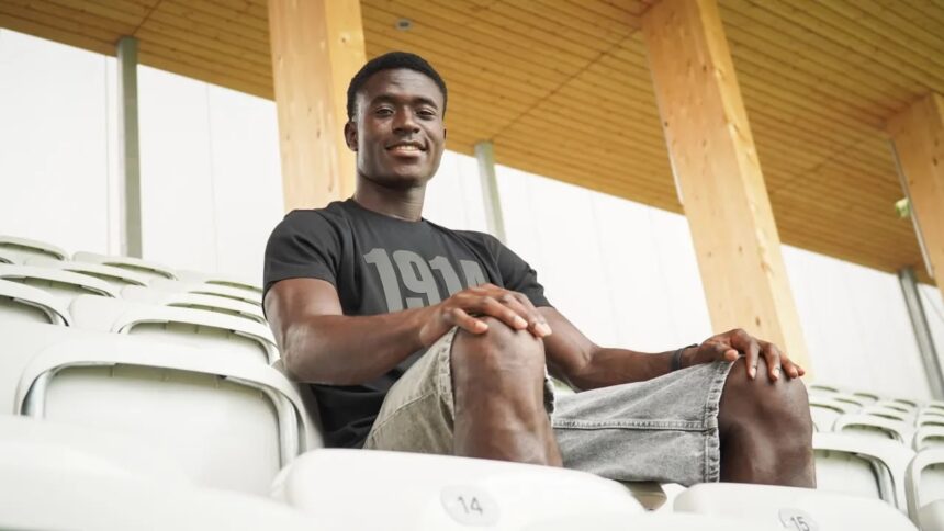Young man sitting in empty stadium seats smiling at the camera wearing a black t shirt and gray shorts with a wooden roof behind him  trybalnews
