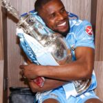 Athlete in a light blue sports kit hugs a large silver trophy in a locker room smiling  trybalnews