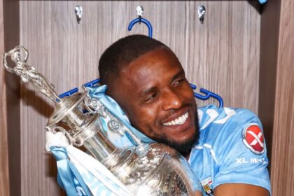 Athlete in a light blue sports kit hugs a large silver trophy in a locker room, smiling.