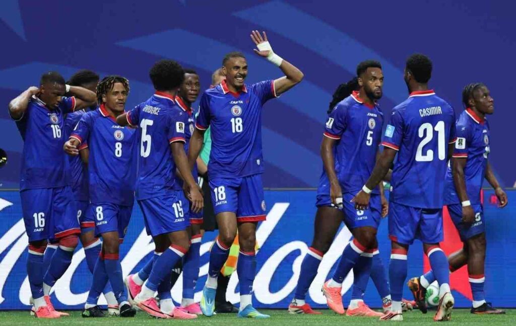 Wilson Isidor Trybal News Haiti forward Wilson Isidor wearing royal blue 18 jersey with red collar and white numbers waves with right hand smiling with mouth open Teammates including 21 CASIMIR 5 15 PROVIDENCE 8 and 16 gather around him on the pitch All wear blue kits with red trim and blue shorts Blurred blue advertising board and green grass in background during FIFA World Cup 2026 Group C match between Morocco and Haiti at Atlanta Stadium Atlanta Georgia on June 24 2026 trybalnews