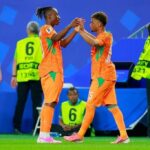 Two soccer players in orange kits share a high five on the field as staff and security stand in the background in a blue stadium setting  trybalnews