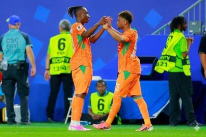 Two soccer players in orange kits share a high-five on the field as staff and security stand in the background in a blue stadium setting.