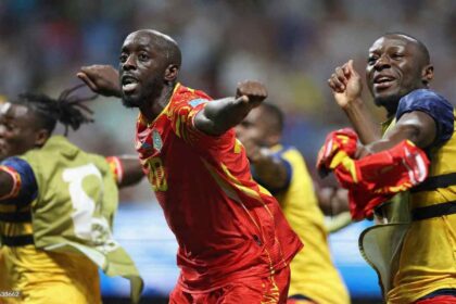 DR Congo forward Yoane Wissa, wearing a red and yellow kit with number 20, celebrates with arms outstretched and fist clenched after scoring. Teammates in yellow and blue training bibs celebrate around him. The 2026 FIFA World Cup Group K match between DR Congo and Uzbekistan took place on June 27, 2026 at Atlanta Stadium in Atlanta, Georgia. A blurred stadium crowd is visible in the background.