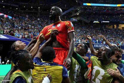 Congo DR forward Yoane Wissa, wearing a red kit with yellow details and number 2, is lifted in celebration by teammates after scoring. Teammates in yellow training bibs over blue kits surround him with arms raised, cheering on the pitch. The 2026 FIFA World Cup Group K match between Congo DR and Uzbekistan took place on June 27, 2026 at Atlanta Stadium in Atlanta, Georgia. A packed stadium crowd and "GOAL" on the stadium screen are visible in the background.
