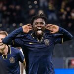 Soccer player in a navy kit celebrates with hands cupping the ears and tongue out on the field  trybalnews