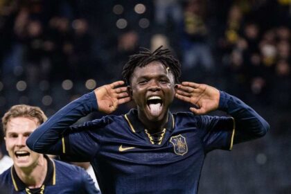 Soccer player in a navy kit celebrates with hands cupping the ears and tongue out on the field.