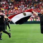 Egypt head coach Hossam Hassan and a staff member run across the green pitch at BC Place holding a large Egyptian flag between them Both wear black Egypt tracksuits Hassan is bald and on the left smiling with a clenched fist The staff member on the right holds a phone Behind them a stadium full of fans in red celebrates Egypts first World Cup win  trybalnews