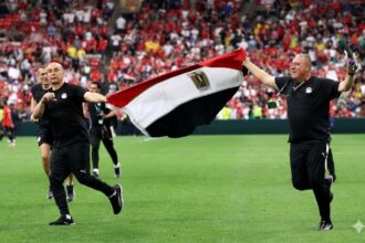 Egypt head coach Hossam Hassan and a staff member run across the green pitch at BC Place holding a large Egyptian flag between them. Both wear black Egypt tracksuits. Hassan is bald and on the left, smiling with a clenched fist. The staff member on the right holds a phone. Behind them, a stadium full of fans in red celebrates Egypt’s first World Cup win.