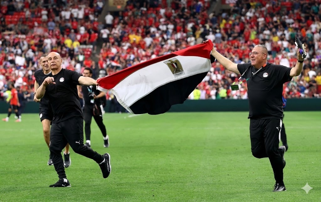 Egypt head coach Hossam Hassan and a staff member run across the green pitch at BC Place holding a large Egyptian flag between them Both wear black Egypt tracksuits Hassan is bald and on the left smiling with a clenched fist The staff member on the right holds a phone Behind them a stadium full of fans in red celebrates Egypts first World Cup win  trybalnews