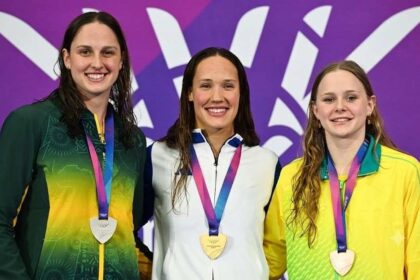 Scottish swimmer Angharad Evans stands in the center of the podium wearing a white, blue and purple jacket and a gold medal, smiling. She is flanked by a South African swimmer in green and yellow on the left with a silver medal, and an Australian swimmer in yellow and green on the right with a bronze medal. The background is purple with the Glasgow 2026 Commonwealth Games logo.