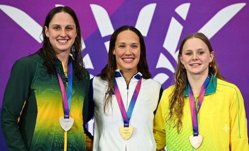 Scottish swimmer Angharad Evans stands in the center of the podium wearing a white blue and purple jacket and a gold medal smiling She is flanked by a South African swimmer in green and yellow on the left with a silver medal and an Australian swimmer in yellow and green on the right with a bronze medal The background is purple with the Glasgow 2026 Commonwealth Games logo  trybalnews