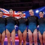 Five gold medalists from Team Australias womens artistic gymnastics team stand together smiling holding the Australian flag behind them Georgia Godwin Kate McDonald Ruby Pass Breanna Scott and Emily Whitehead wear matching blue and black leotards with AUS on them The background shows The Arena in Glasgow during the 2026 Commonwealth Games on July 25 2026  trybalnews
