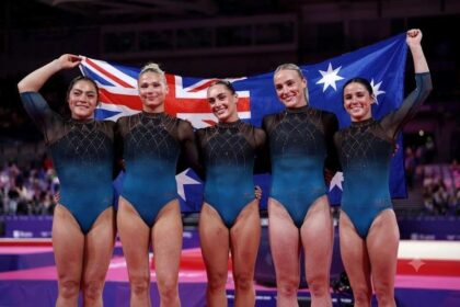 Five gold medalists from Team Australia’s women’s artistic gymnastics team stand together smiling, holding the Australian flag behind them. Georgia Godwin, Kate McDonald, Ruby Pass, Breanna Scott and Emily Whitehead wear matching blue and black leotards with "AUS" on them. The background shows The Arena in Glasgow during the 2026 Commonwealth Games on July 25, 2026.