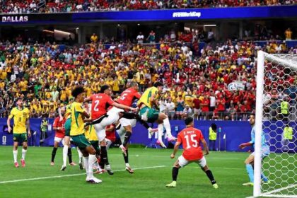Mohamed Hany of Egypt, wearing a red jersey with number 2, attempts a defensive clearance in front of goal during the FIFA World Cup 2026 Round of 32 match between Australia and Egypt at Dallas Stadium in Arlington, Texas, on July 3, 2026. The ball is in the air near the goal line as Australian players in yellow and green and other Egyptian players in red contest a header. The score was leveled at 1-1 following an own goal. A packed stadium crowd fills the background with "DALLAS" and "#FIFAWorldCup" visible on the stadium signage.