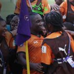 Group of soccer players in orange and brown uniforms celebrate with arms around each other a purple flag in the foreground  trybalnews