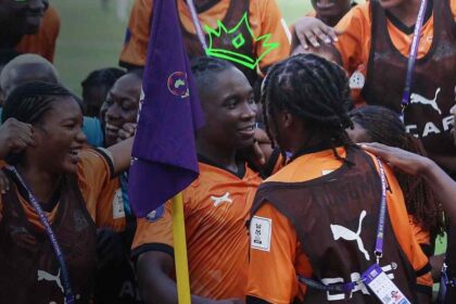 Group of soccer players in orange and brown uniforms celebrate with arms around each other, a purple flag in the foreground.