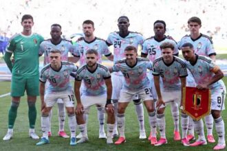 Belgium national team players pose for a team photo before the FIFA World Cup 2026 Round of 16 match against USA at Seattle Stadium in Seattle on July 6, 2026. The goalkeeper wears a green kit while outfield players wear light blue and pink patterned away jerseys with white shorts. A player holds a red Belgium pennant.
