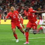 Switzerland forward Breel Embolo wearing a red number 7 jersey and pink cleats celebrates mid air with his right arm outstretched after scoring his teams first goal Teammate wearing number 11 in a matching red Switzerland kit runs beside him The action is on a green pitch at BC Place Stadium with a blurred crowd and yellow Lays advertising boards in the background during the FIFA World Cup 2026 Round of 32 match against Algeria  trybalnews