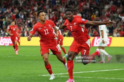 Switzerland forward Breel Embolo, wearing a red number 7 jersey and pink cleats, celebrates mid-air with his right arm outstretched after scoring his team’s first goal. Teammate wearing number 11 in a matching red Switzerland kit runs beside him. The action is on a green pitch at BC Place Stadium with a blurred crowd and yellow Lay’s advertising boards in the background during the FIFA World Cup 2026 Round of 32 match against Algeria.