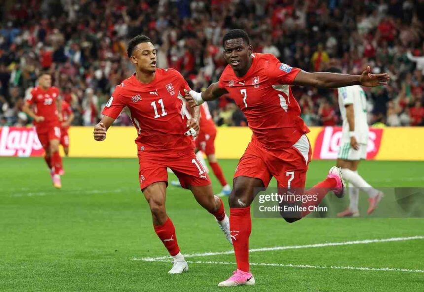 Switzerland forward Breel Embolo wearing a red number 7 jersey and pink cleats celebrates mid air with his right arm outstretched after scoring his teams first goal Teammate wearing number 11 in a matching red Switzerland kit runs beside him The action is on a green pitch at BC Place Stadium with a blurred crowd and yellow Lays advertising boards in the background during the FIFA World Cup 2026 Round of 32 match against Algeria  trybalnews