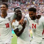 Three England footballers celebrate a goal on the pitch arms around each other amid cheering fans in the stands in white kits with red numbers 10 and 24 for context  trybalnews
