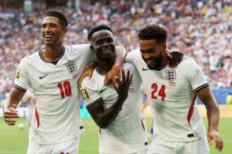 Three England footballers celebrate a goal on the pitch, arms around each other, amid cheering fans in the stands in white kits with red numbers 10 and 24 for context.