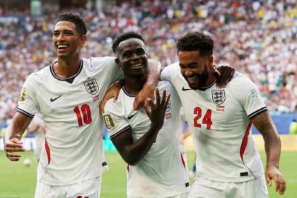 Three England footballers celebrate a goal on the pitch, arms around each other, amid cheering fans in the stands in white kits with red numbers 10 and 24 for context.