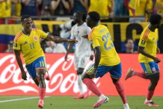 Jhon Arias of Colombia, wearing a yellow jersey with number 11 and blue shorts, celebrates with a wide smile and outstretched arm after scoring his team's first goal. He is joined by teammate Davinson Sanchez, defender #23, also in a yellow jersey and blue shorts, running toward him on the pitch. Another Colombian player in yellow is visible to the right. In the background, a Ghana player in a white kit walks away, with Colombian fans in yellow shirts and a red Coca-Cola advertising board behind them. The scene is from the 2026 World Cup Round of 32 match between Colombia and Ghana at Kansas City Stadium on July 3, 2026.