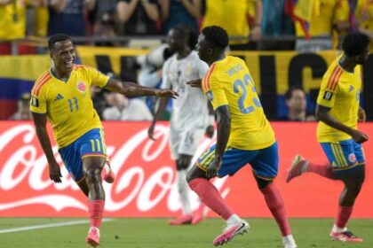 Jhon Arias of Colombia, wearing a yellow jersey with number 11 and blue shorts, celebrates with a wide smile and outstretched arm after scoring his team's first goal. He is joined by teammate Davinson Sanchez, defender #23, also in a yellow jersey and blue shorts, running toward him on the pitch. Another Colombian player in yellow is visible to the right. In the background, a Ghana player in a white kit walks away, with Colombian fans in yellow shirts and a red Coca-Cola advertising board behind them. The scene is from the 2026 World Cup Round of 32 match between Colombia and Ghana at Kansas City Stadium on July 3, 2026.