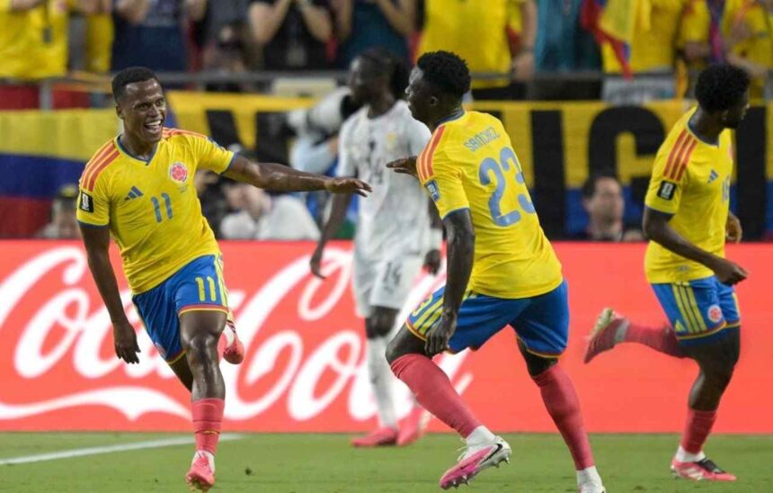 Jhon Arias of Colombia wearing a yellow jersey with number 11 and blue shorts celebrates with a wide smile and outstretched arm after scoring his teams first goal He is joined by teammate Davinson Sanchez defender 23 also in a yellow jersey and blue shorts running toward him on the pitch Another Colombian player in yellow is visible to the right In the background a Ghana player in a white kit walks away with Colombian fans in yellow shirts and a red Coca Cola advertising board behind them The scene is from the 2026 World Cup Round of 32 match between Colombia and Ghana at Kansas City Stadium on July 3 2026  trybalnews