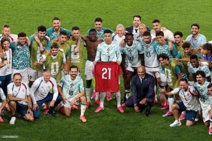 Portugal’s Cristiano Ronaldo, wearing a light blue and white number 7 jersey, stands center holding up a red Portugal jersey with the number 21 to commemorate late teammate Diogo Jota. He is surrounded by smiling teammates and coaching staff in matching kits and training bibs, posing together on a green pitch after their 2-1 FIFA World Cup 2026 Round of 32 win over Croatia in Toronto.